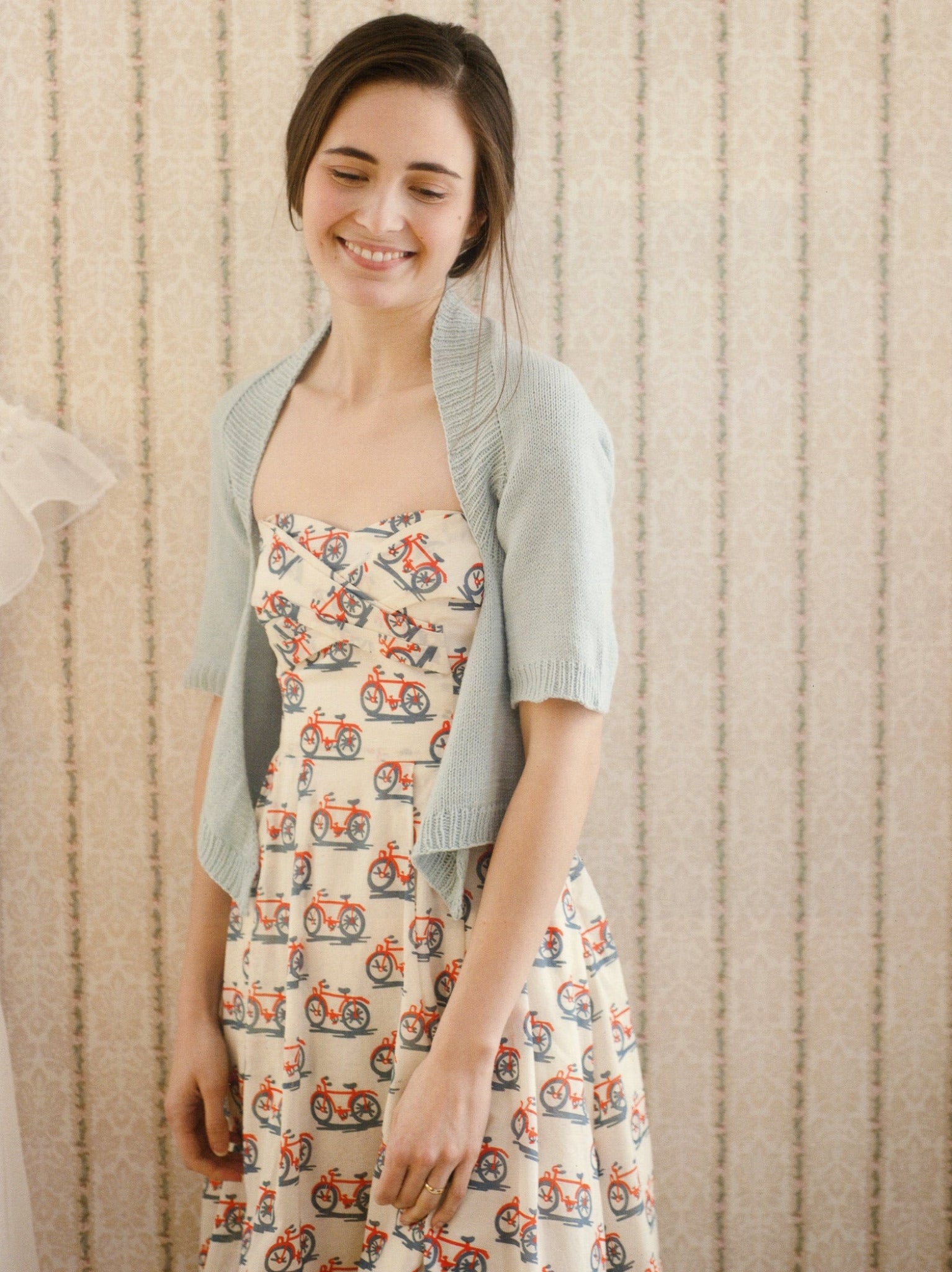 A young woman with long dark hair and a content expression is wearing the Knitbot Essentials by Never Not Knitting - a light blue shrug over a sleeveless dress adorned with a popular pattern of red bicycles. The background features a light, patterned wallpaper, suggesting she is indoors.