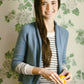 A woman with long brown hair, wearing a blue cardigan from Never Not Knitting's Knitbot Essentials over a striped shirt, smiles while holding an orange. The background features a floral wallpaper with green leaves and white flowers.