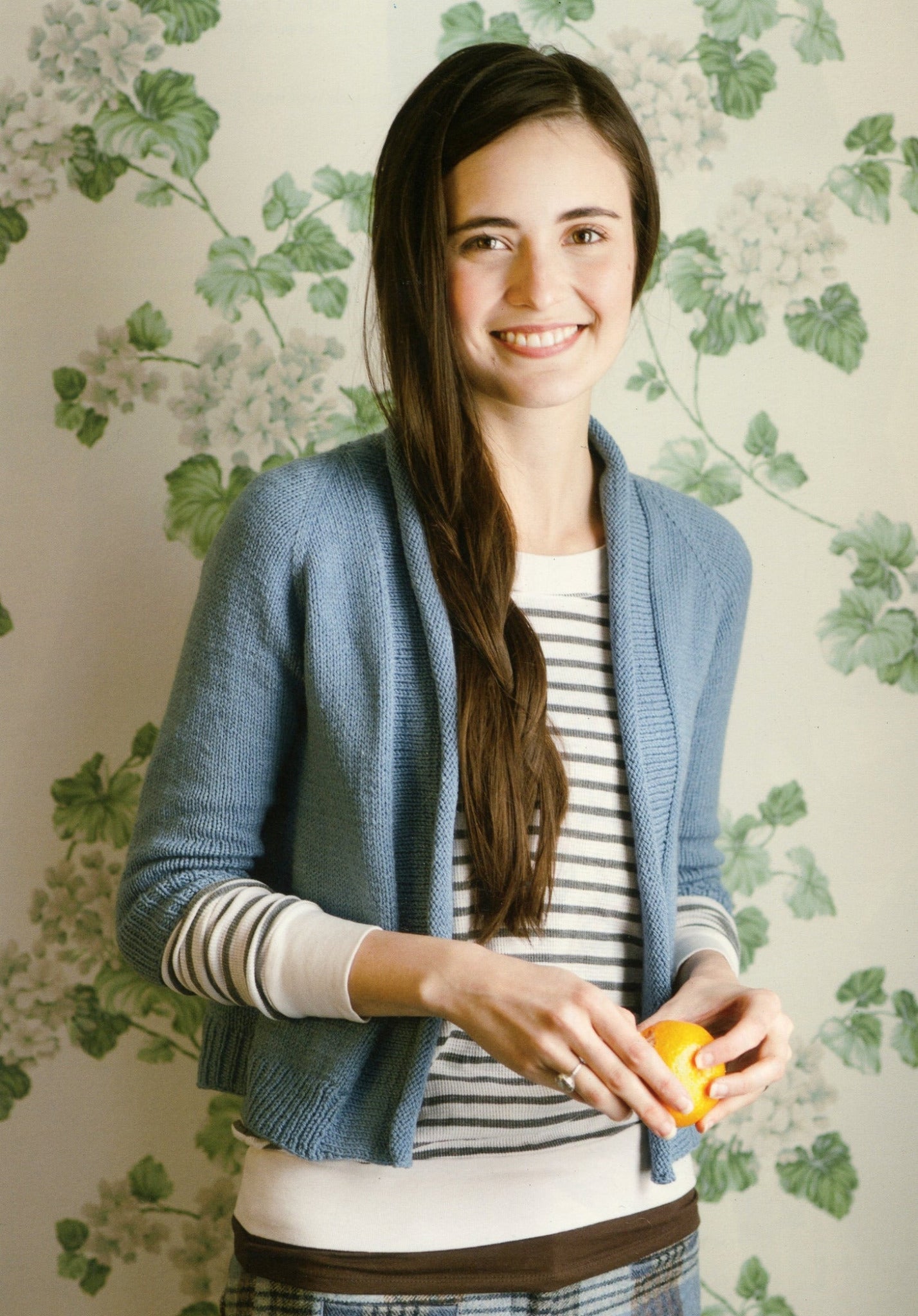 A woman with long brown hair, wearing a blue cardigan from Never Not Knitting's Knitbot Essentials over a striped shirt, smiles while holding an orange. The background features a floral wallpaper with green leaves and white flowers.