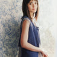 A woman with straight brown hair gazes at the camera. She wears a sleeveless blue vest over a light gray dress reminiscent of delicate Knitbot Linen patterns by Never Not Knitting. The background features a blue and white floral wallpaper. She stands with one hand resting on a wicker chair. Natural light illuminates the scene.