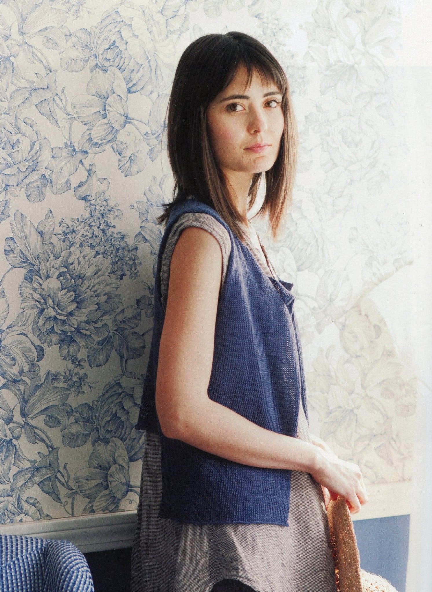 A woman with straight brown hair gazes at the camera. She wears a sleeveless blue vest over a light gray dress reminiscent of delicate Knitbot Linen patterns by Never Not Knitting. The background features a blue and white floral wallpaper. She stands with one hand resting on a wicker chair. Natural light illuminates the scene.