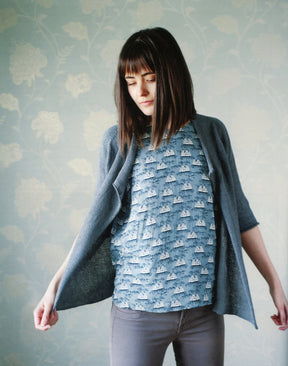 A woman with shoulder-length brown hair stands against a floral-patterned wallpaper. She is wearing a blue cardigan, crafted from Never Not Knitting's Knitbot Linen, over a light blue top with a sailboat print and gray pants. She looks down while holding the edges of her cozy knit cardigan open.