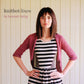 A woman stands confidently with hands on hips, wearing a black and white striped dress and a pink knit cardigan. The background features vertical white paneling. Text reads, "Knitbot Linen by Never Not Knitting," showcasing elegant patterns that highlight the versatility of knits in linen.