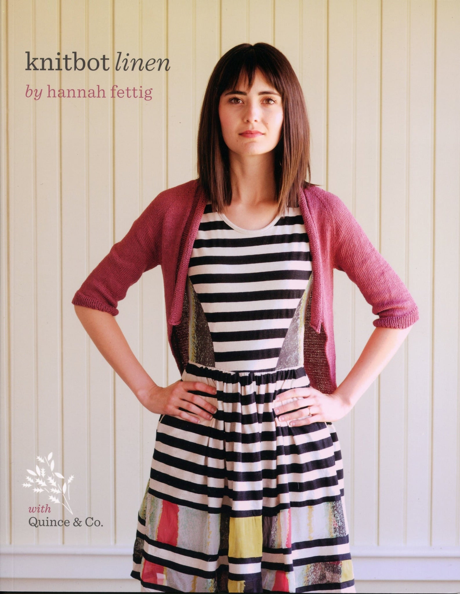 A woman with straight dark hair stands against a light-colored, vertical wooden wall. She wears a pink knit cardigan over a sleeveless black-and-white striped dress with colorful abstract patterns near the hem. Text on the image reads, "Knitbot Linen by Never Not Knitting, featuring Halcyon Yarn Block Island Blend.