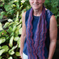 An older adult with short gray hair and glasses stands outdoors near a garden. They wear a blue sleeveless shirt, white pants, and a purple wavy-patterned scarf from the "Madder - Anthology 1 / Camilla, Sibella, Imogen, & Beatrice" collection by Never Not Knitting. The background features green plants and leaves, creating a natural, serene setting.