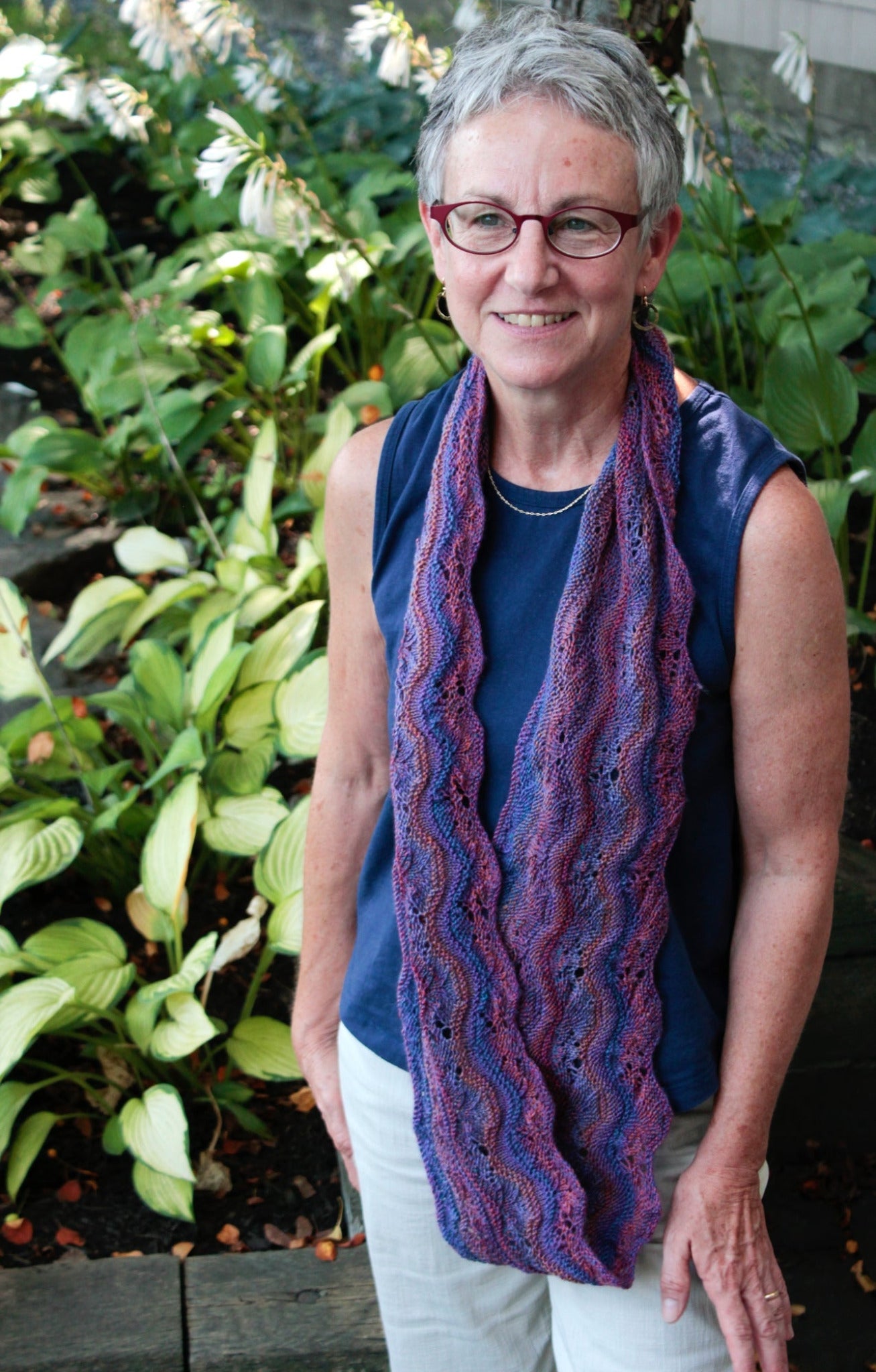 An older adult with short gray hair and glasses stands outdoors near a garden. They wear a blue sleeveless shirt, white pants, and a purple wavy-patterned scarf from the "Madder - Anthology 1 / Camilla, Sibella, Imogen, & Beatrice" collection by Never Not Knitting. The background features green plants and leaves, creating a natural, serene setting.