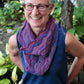 A person with short white hair and glasses smiles warmly while standing outside. They are wearing a sleeveless blue top paired with a purple, wavy patterned scarf from Never Not Knitting's Madder - Anthology 1, featuring the designs Camilla, Sibella, Imogen, & Beatrice. The background features an array of green leafy plants, creating a serene atmosphere.