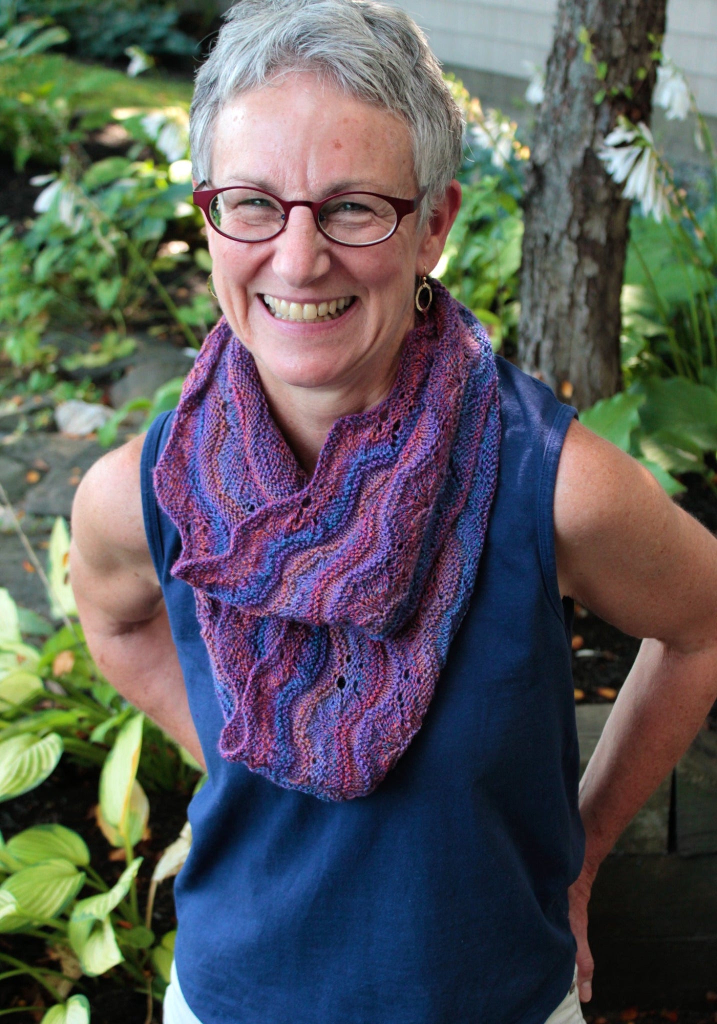 A person with short white hair and glasses smiles warmly while standing outside. They are wearing a sleeveless blue top paired with a purple, wavy patterned scarf from Never Not Knitting's Madder - Anthology 1, featuring the designs Camilla, Sibella, Imogen, & Beatrice. The background features an array of green leafy plants, creating a serene atmosphere.