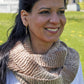 A woman with long dark hair smiles while standing outdoors on a sunny day. She is wearing a hand-knitted, multi-toned scarf from the Madder - Anthology 2 / Simple Pleasures collection by Never Not Knitting, which highlights her love for unique accessories. The scarf is paired with a light-colored top. The background features a grassy area with trees and a white fence.