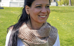 A woman with long dark hair smiles while standing outdoors on a sunny day. She is wearing a hand-knitted, multi-toned scarf from the Madder - Anthology 2 / Simple Pleasures collection by Never Not Knitting, which highlights her love for unique accessories. The scarf is paired with a light-colored top. The background features a grassy area with trees and a white fence.