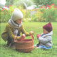 Two young children, both wearing cozy sweaters from the "A Sense of Place" collection by Never Not Knitting, sit on a grassy lawn. The older child, kneeling, places apples into a wooden basket while the younger child, sitting, holds and examines an apple. Red flowers bloom in the background.