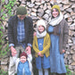 A family of four, warmly dressed in the "A Sense of Place" knit hats and cardigans from Never Not Knitting, stands in front of a stacked woodpile. The father cradles a smiling baby adorned in a blue knitted hat and shirt. A young girl, wearing a yellow cardigan and green hat from the same collection, stands between her parents. The mother is outfitted in a matching gray hat and cardigan from Never Not Knitting's "A Sense of Place.