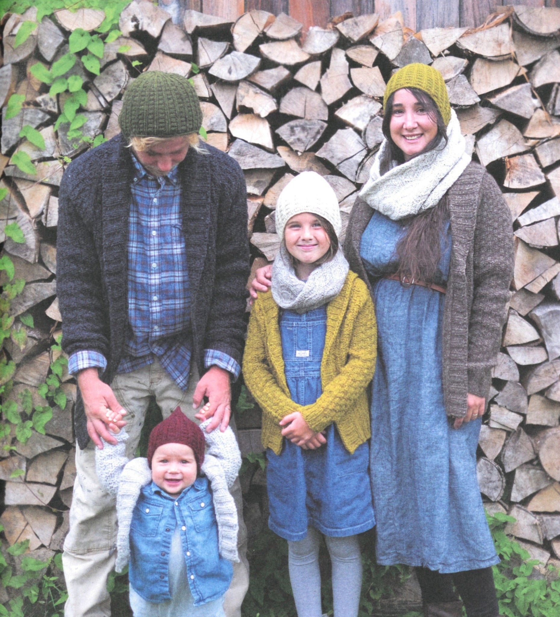 A family of four, warmly dressed in the "A Sense of Place" knit hats and cardigans from Never Not Knitting, stands in front of a stacked woodpile. The father cradles a smiling baby adorned in a blue knitted hat and shirt. A young girl, wearing a yellow cardigan and green hat from the same collection, stands between her parents. The mother is outfitted in a matching gray hat and cardigan from Never Not Knitting's "A Sense of Place.