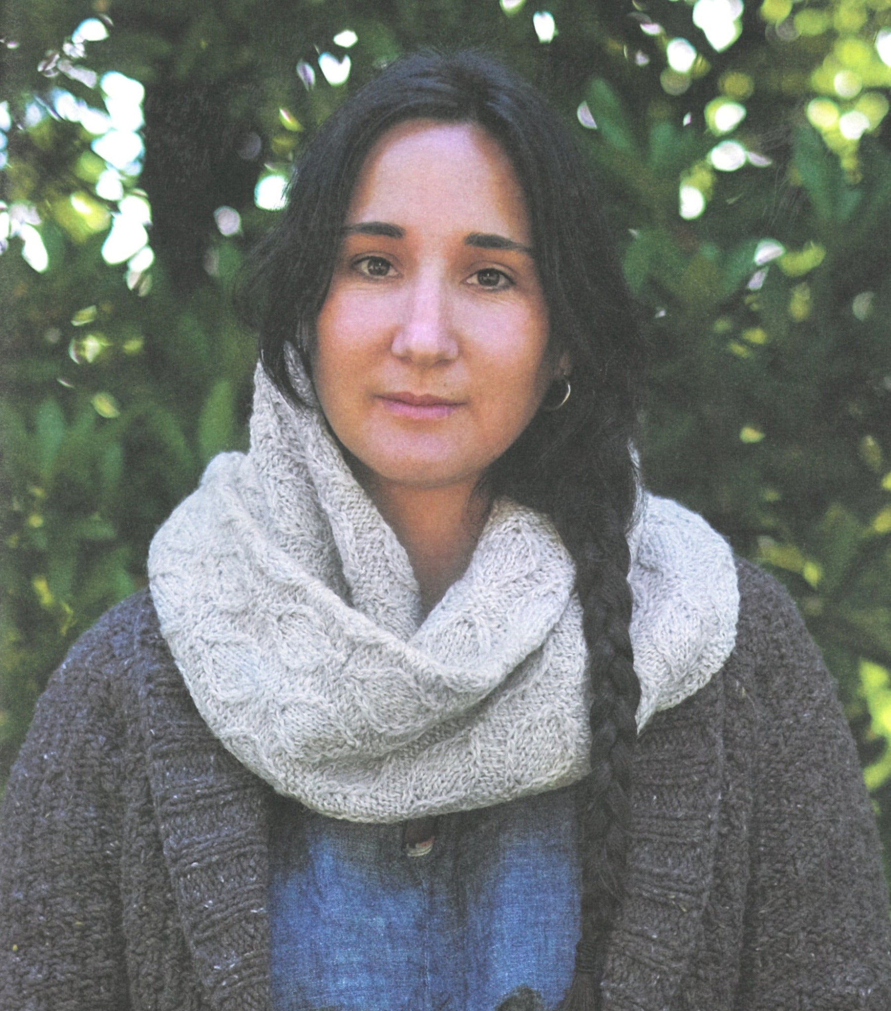 A person with long dark hair braided over one shoulder wears "A Sense of Place," a thick, light-colored knit scarf adorned with intricate wooly knitting patterns from Never Not Knitting, and a dark sweater. They stand against a backdrop of lush green foliage, their expression calm and serene.