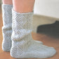 Close-up view of a person's feet donning "A Sense of Place" socks by Never Not Knitting. The light gray, leaf-like textured pattern and wooly knitting make the socks seem incredibly soft and cozy, extending up to mid-calf. The person is indoors on a wooden floor, with some furniture partially visible in the background.