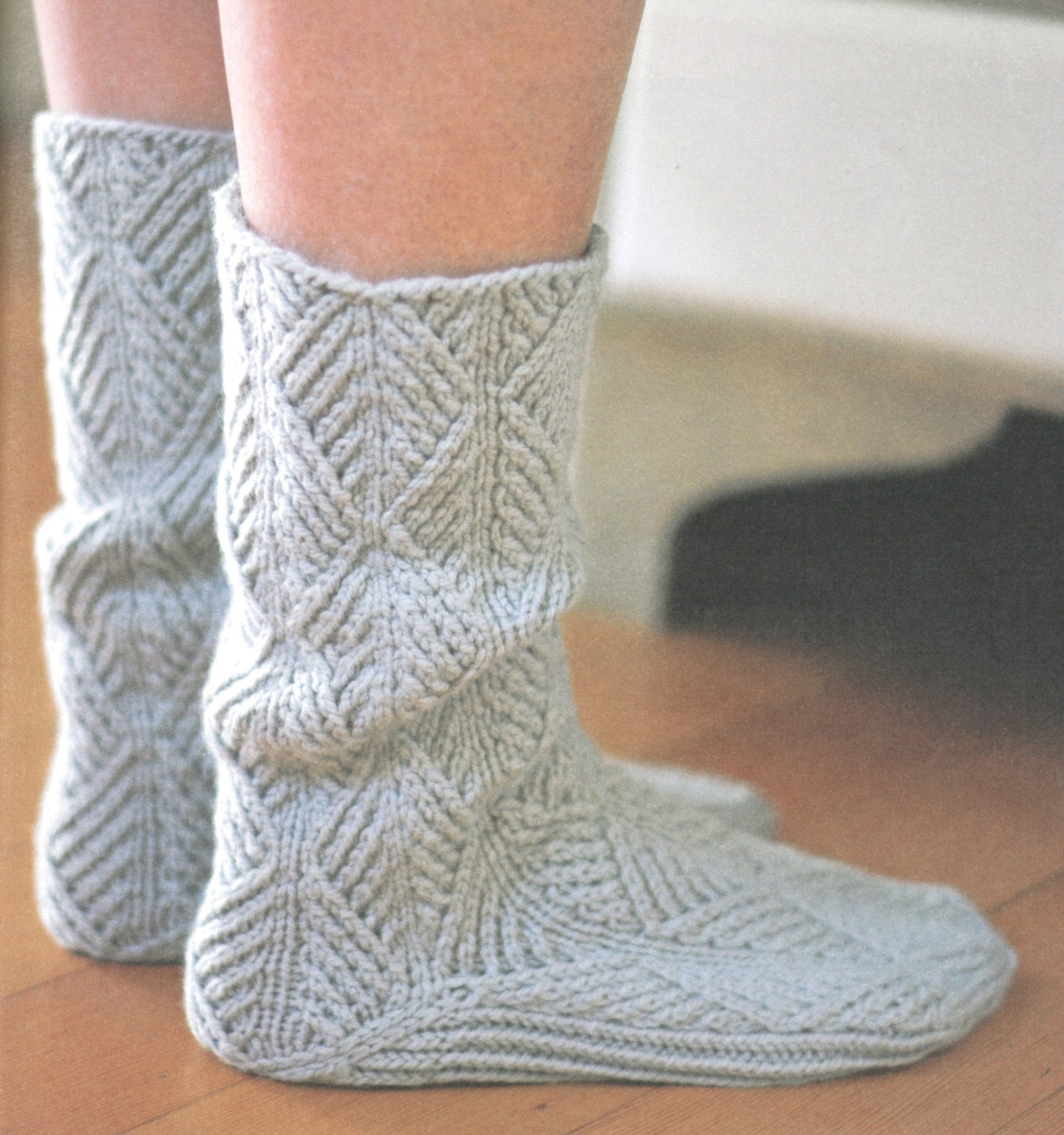 Close-up view of a person's feet donning "A Sense of Place" socks by Never Not Knitting. The light gray, leaf-like textured pattern and wooly knitting make the socks seem incredibly soft and cozy, extending up to mid-calf. The person is indoors on a wooden floor, with some furniture partially visible in the background.