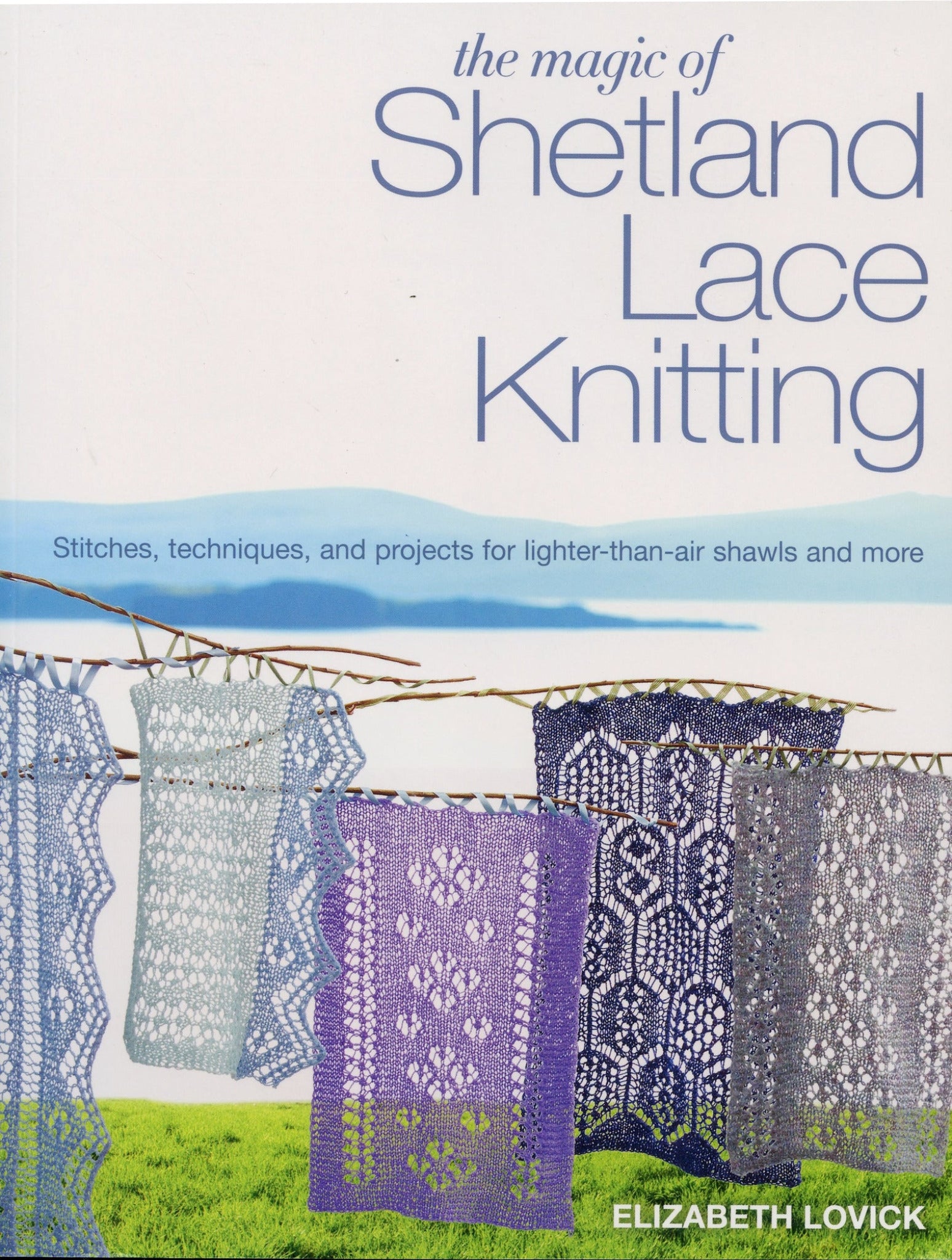 The book cover of "The Magic of Shetland Lace Knitting" by MacMillan/MPS Holdings LLC, designed by Elizabeth Lovick, showcases intricate lace patterns in hues of white, lavender, and gray. These beautiful designs are displayed on a clothesline set against a picturesque grassy field with a clear blue sky. The subtext: "Stitches, techniques, and projects for lighter-than-air shawls and more," makes it an ideal choice for beginner knitters.