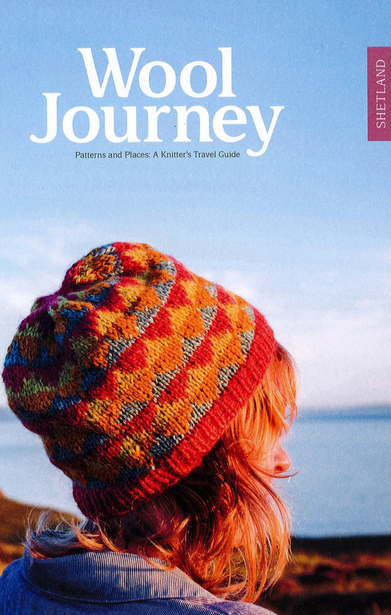 Cover of "Wool Journey: Shetland" by Pom Pom Publishing. Features a person with shoulder-length hair wearing a colorful knitted hat. The background displays a serene coastal landscape with a clear blue sky. Text: "Wool Journey: Shetland – A Knitter's Travel Guide, offering unique knitting patterns inspired by Shetland.”