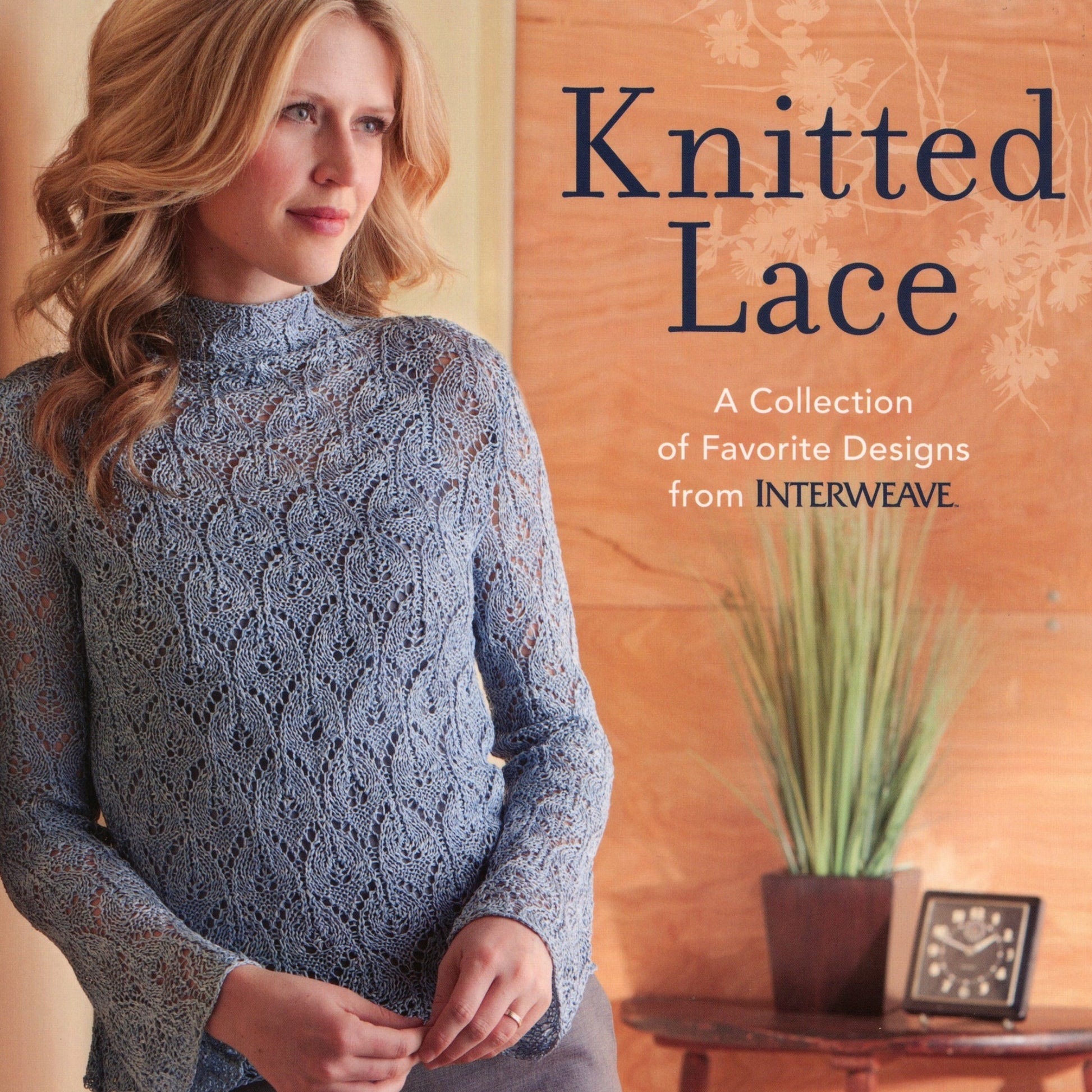 A woman with wavy blonde hair is wearing a light blue lace-knit sweater. She stands with her hands crossed in front, gazing to the side. Next to her, the text reads "Knitted Lace by Ingram Content," celebrating lace projects and intricate lace charts. A potted plant sits on a table behind her.