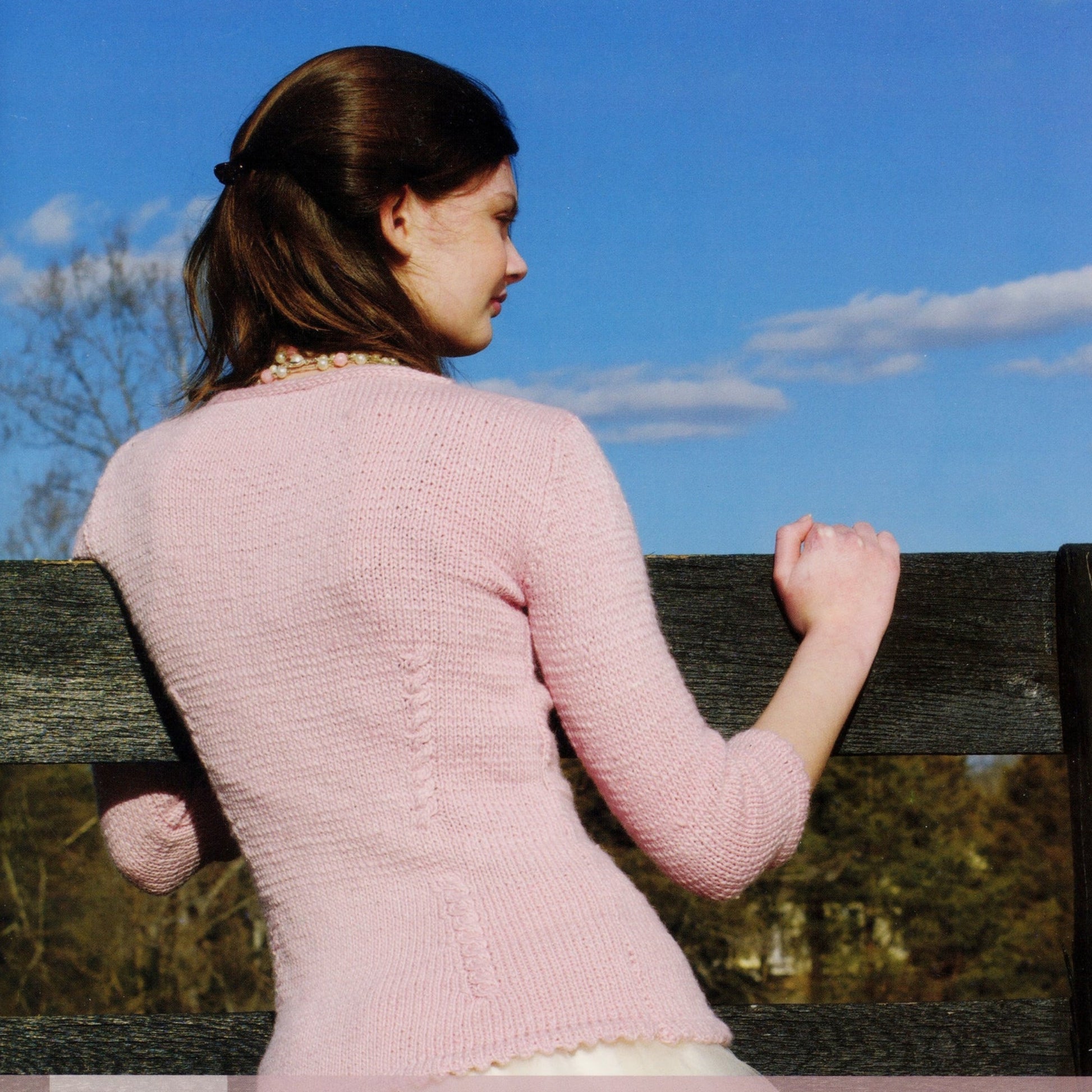 A woman with brown hair, wearing a pink long-sleeve sweater from the "Moonshine - 14 Winsome Patterns" collection by Knitting Fever / Euro Yarns and a beaded necklace, leans against a wooden fence with her right hand. She is outdoors on a sunny day under a clear blue sky at Juniper Moon Farm.