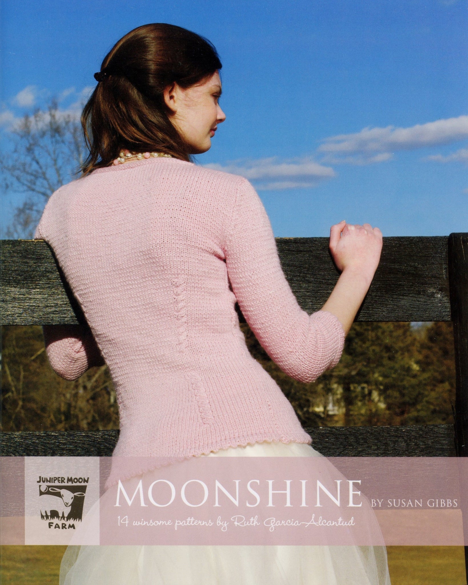 A woman with brown hair in a ponytail, wearing a light pink knitted sweater and white skirt, leans against a wooden fence under a clear blue sky. The text at the bottom reads "Moonshine - 14 Winsome Patterns" by Ruth Garcia-Alcántud from Juniper Moon Farm, featuring luxurious blends by Knitting Fever / Euro Yarns.