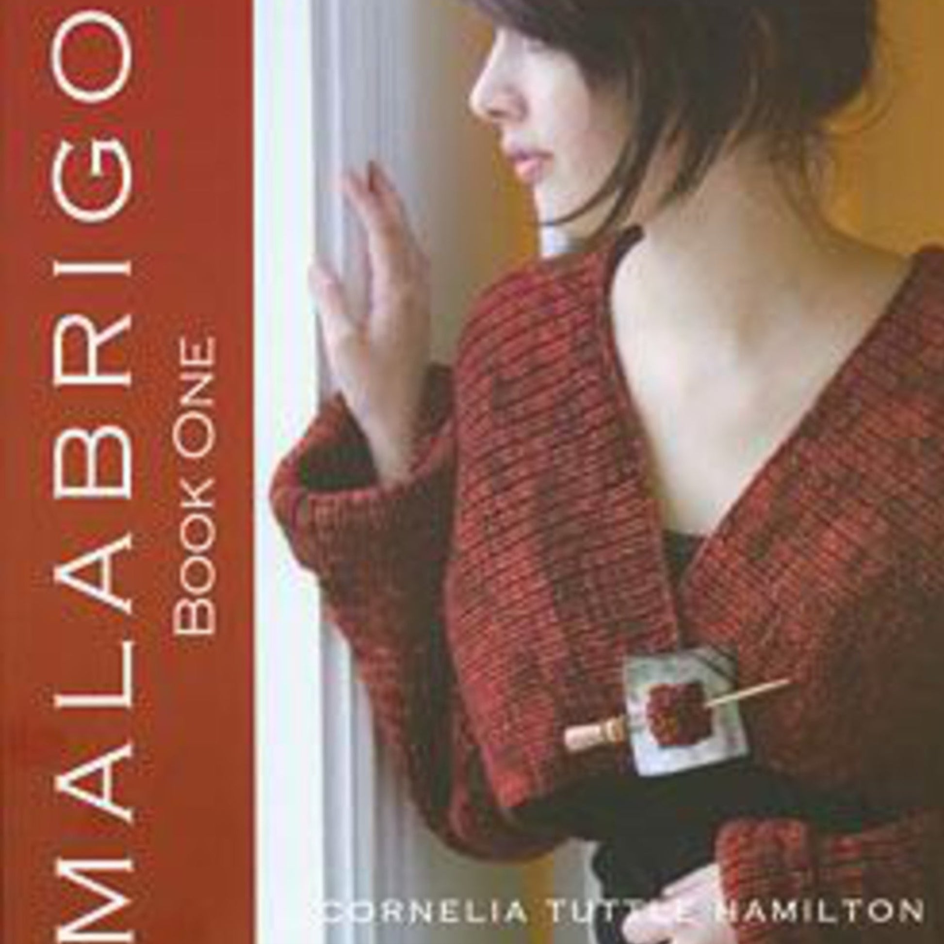 A woman with dark hair stands near a white wall, wearing a textured red sweater with a black belt. She is looking to the side with her hand resting against the wall. The cover of Malabrigo Book One by Malabrigo Yarn, featuring designs by Cornelia Tuttle Hamilton, showcases scarves and sweaters.