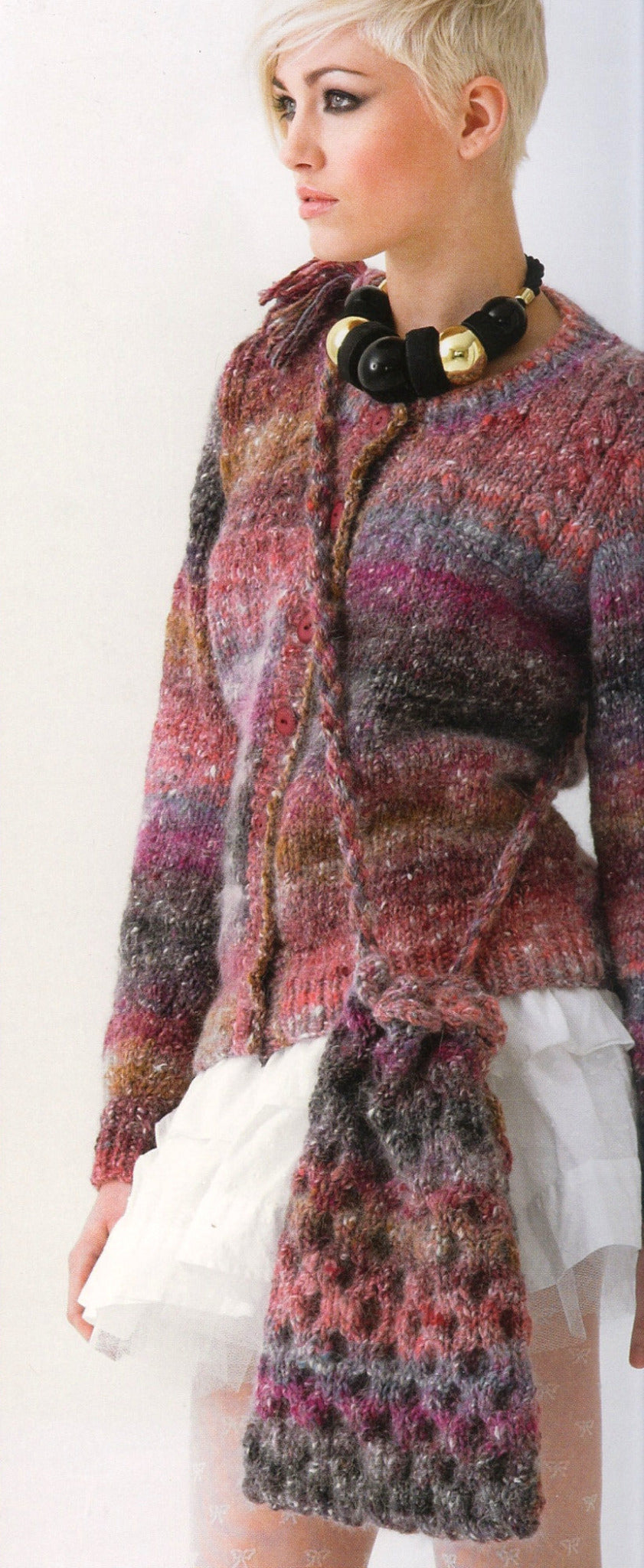 A person with short, platinum blonde hair is wearing trendy knitwear from the product line Noro Now! by Knitting Fever / Euro Yarns: a multicolored cardigan and scarf showcasing colorful knitting patterns in shades of pink, purple, gray, and brown. They also have on a black necklace with large, spherical beads and a layered white tulle skirt.