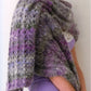 A person with short, blonde hair is wearing a light purple sleeveless top and a textured, multicolored purple and gray shawl from the Noro Now! collection by Knitting Fever / Euro Yarns draped over their shoulders. Showcasing trendy knitwear, they are looking to the side, with part of a mauve-colored ruffled skirt visible at the bottom.