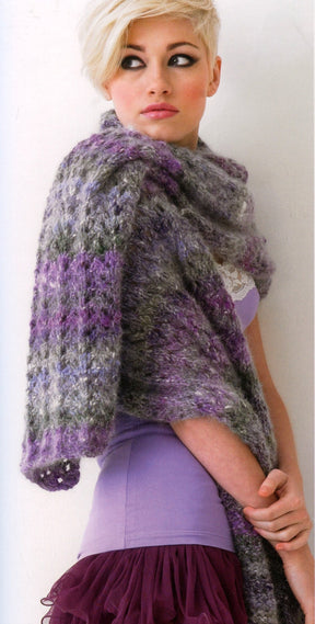 A person with short, blonde hair is wearing a light purple sleeveless top and a textured, multicolored purple and gray shawl from the Noro Now! collection by Knitting Fever / Euro Yarns draped over their shoulders. Showcasing trendy knitwear, they are looking to the side, with part of a mauve-colored ruffled skirt visible at the bottom.