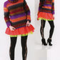 Two models showcasing colorful winter fashion from Noro Now! by Knitting Fever / Euro Yarns. Both are dressed in trendy knitwear, including multicolored striped sweaters, neon tutus, black leggings with lace patterns on the side, and black ankle boots. One model sports a short pixie haircut, while the other complements her look with a matching knit hat.