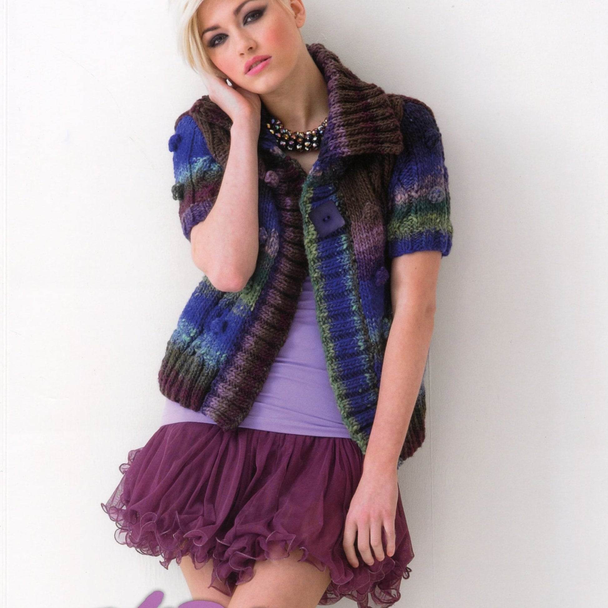 A woman with blonde hair, wearing stylish pieces from the Noro Now! collection by Knitting Fever / Euro Yarns, including a multi-colored knit cardigan with large buttons, a purple top, a statement necklace, and a ruffled purple skirt stands against a white wall. She rests her head on her right hand and gazes into the distance.