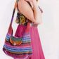 A person dressed in a colorful sundress is carrying a vibrant, multicolored knit shoulder bag crafted from Noro Spring into Summer yarn by Knitting Fever / Euro Yarns, complete with a button closure. The sundress boasts a smocked floral top and a bright pink skirt. The person's face is not visible in the image.