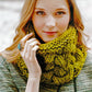 A young woman with long, wavy, auburn hair gazes into the camera. She is wearing a textured gray sweater and a chunky, green knit scarf made from Malabrigo Yarn featured in Malabrigo Book 5 - In Soho. Her hand, adorned with a large oval turquoise ring, gently touches the sophisticated cowl near her face.