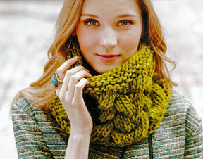 A young woman with long, wavy, auburn hair gazes into the camera. She is wearing a textured gray sweater and a chunky, green knit scarf made from Malabrigo Yarn featured in Malabrigo Book 5 - In Soho. Her hand, adorned with a large oval turquoise ring, gently touches the sophisticated cowl near her face.