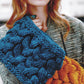 A person with long hair stands holding two sophisticated cowls from the Malabrigo Book 5 - In Soho collection by Malabrigo Yarn: one in blue and the other in orange. They are wearing a textured jacket, and their expression is neutral.