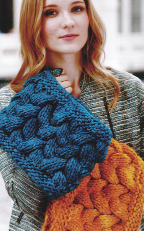 A person with long hair stands holding two sophisticated cowls from the Malabrigo Book 5 - In Soho collection by Malabrigo Yarn: one in blue and the other in orange. They are wearing a textured jacket, and their expression is neutral.