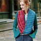 A woman with light hair is wearing a teal blazer and a textured knitted scarf made with yarn from Malabrigo Yarn's "Malabrigo Book 5 - In Soho" in red and blue. She stands outdoors with her hands in her pockets, looking to the side, in front of a building and an empty street.