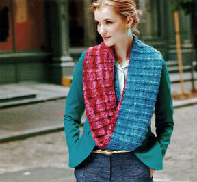 A woman with light hair is wearing a teal blazer and a textured knitted scarf made with yarn from Malabrigo Yarn's "Malabrigo Book 5 - In Soho" in red and blue. She stands outdoors with her hands in her pockets, looking to the side, in front of a building and an empty street.