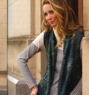 A woman with long, wavy hair stands by a stone wall, looking at the camera with a slight smile. She is wearing a striped long-sleeve top and a blue-green knitted vest made from soft Merino yarns featured in Malabrigo Book 4 by Malabrigo Yarn, with one hand placed on her hip. The background features a stone building with a window.