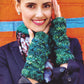 A person smiles at the camera, wearing a dark blazer and a patterned scarf while showcasing chunky, green and blue hand-knit fingerless gloves made from Malabrigo Yarn's Rasta, as featured in Malabrigo Book 7 - In Brooklyn. They hold their hands up to their face against a vibrant orange and blue wall backdrop.