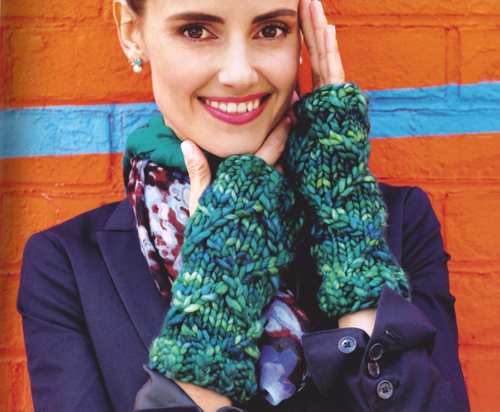 A person smiles at the camera, wearing a dark blazer and a patterned scarf while showcasing chunky, green and blue hand-knit fingerless gloves made from Malabrigo Yarn's Rasta, as featured in Malabrigo Book 7 - In Brooklyn. They hold their hands up to their face against a vibrant orange and blue wall backdrop.