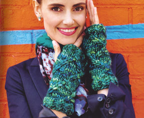 A person smiles at the camera, wearing a dark blazer and a patterned scarf while showcasing chunky, green and blue hand-knit fingerless gloves made from Malabrigo Yarn's Rasta, as featured in Malabrigo Book 7 - In Brooklyn. They hold their hands up to their face against a vibrant orange and blue wall backdrop.