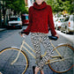 A person stands on a cobblestone street holding a beige bicycle. They are wearing a red knitted sweater with a high collar, crafted from Malabrigo Book 7 - In Brooklyn using Malabrigo Yarn's Rasta, and black-and-white patterned pants. Cars are parked along the sides of the street, and trees line the background.