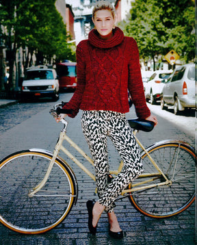 A person stands on a cobblestone street holding a beige bicycle. They are wearing a red knitted sweater with a high collar, crafted from Malabrigo Book 7 - In Brooklyn using Malabrigo Yarn's Rasta, and black-and-white patterned pants. Cars are parked along the sides of the street, and trees line the background.