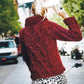 A person with blonde hair tied in a ponytail, wearing a textured red sweater knitted with Malabrigo Yarn from the Malabrigo Book 7 - In Brooklyn, Rasta collection, rides a bicycle down a tree-lined urban street. The back view shows a patterned design on the sweater and the individual's hand adjusting their hair.