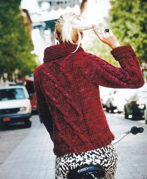 A person with blonde hair tied in a ponytail, wearing a textured red sweater knitted with Malabrigo Yarn from the Malabrigo Book 7 - In Brooklyn, Rasta collection, rides a bicycle down a tree-lined urban street. The back view shows a patterned design on the sweater and the individual's hand adjusting their hair.