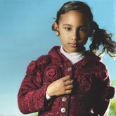 A young girl with dark hair tied in a loose ponytail stands against a blue sky background. She is wearing a maroon knitted sweater featuring rose patterns from Malabrigo Yarn's Malabrigo Book 9 - Niños collection, holding one side of it. She appears contemplative, looking directly into the camera.