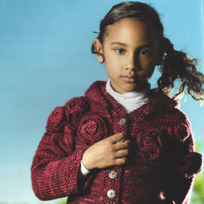 A young girl with dark hair tied in a loose ponytail stands against a blue sky background. She is wearing a maroon knitted sweater featuring rose patterns from Malabrigo Yarn's Malabrigo Book 9 - Niños collection, holding one side of it. She appears contemplative, looking directly into the camera.