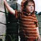 A young child wearing a knitted hoodie with a gradient pattern of red, orange, and brown hues stands outdoors, holding onto a wooden structure. The child looks off into the distance with a serious expression. This unique pattern is one of the highlights from *Malabrigo Book 9 - Niños* by Malabrigo Yarn. Grass and rocks are visible in the background.