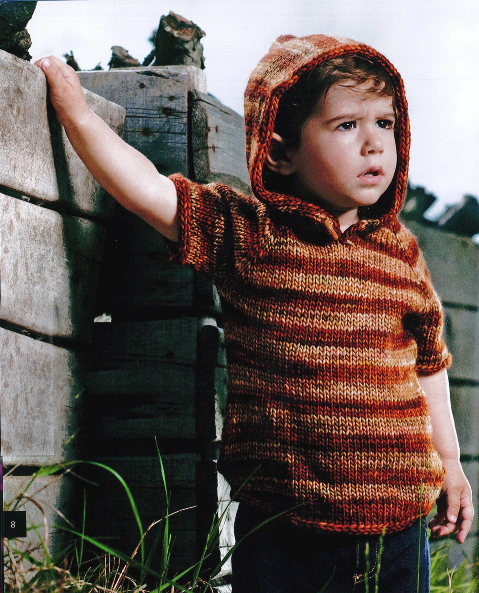 A young child wearing a knitted hoodie with a gradient pattern of red, orange, and brown hues stands outdoors, holding onto a wooden structure. The child looks off into the distance with a serious expression. This unique pattern is one of the highlights from *Malabrigo Book 9 - Niños* by Malabrigo Yarn. Grass and rocks are visible in the background.